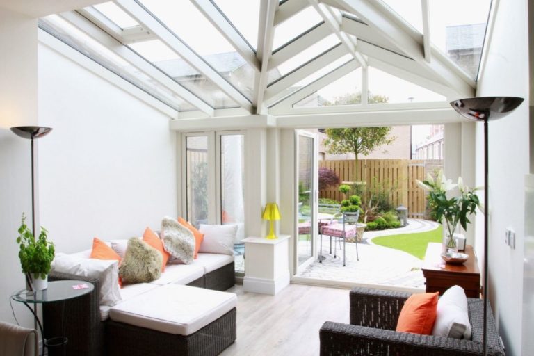 Modern orangery interior featuring white rattan furniture, vibrant cushions, integrated spotlighting, and a view of a landscaped garden.