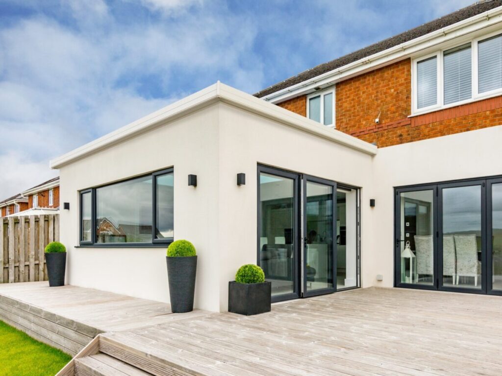 a cream rendered flat roof glass extension onto a brick and cream rendered house with black framed windows and doors