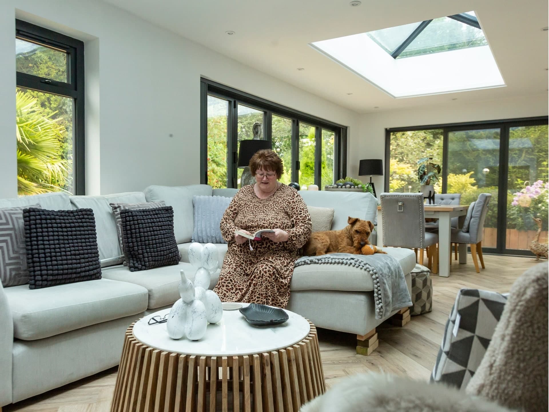 Photo showing a lady reading a book on a cosy sofa next to her dog, in her new modular glass kitchen/diner extension with wooded herringbone flooring and modern grey framed patio doors as well as a sky light .