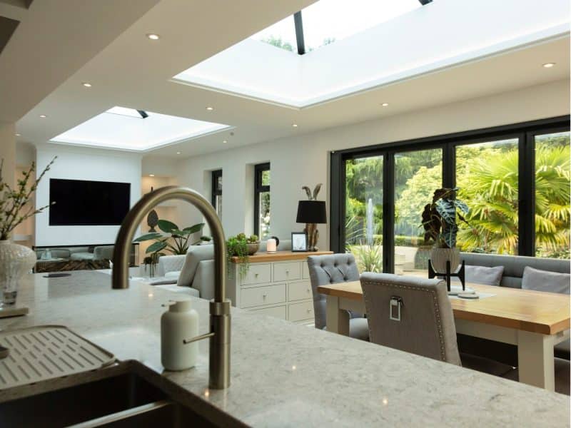 an image showing a new modular glass extension from the kitchen looking across to the dining area and family area with cosy searing and a wall mounted TV. Sky lights flood it with light along with the modern, grey, framed patio doors.
