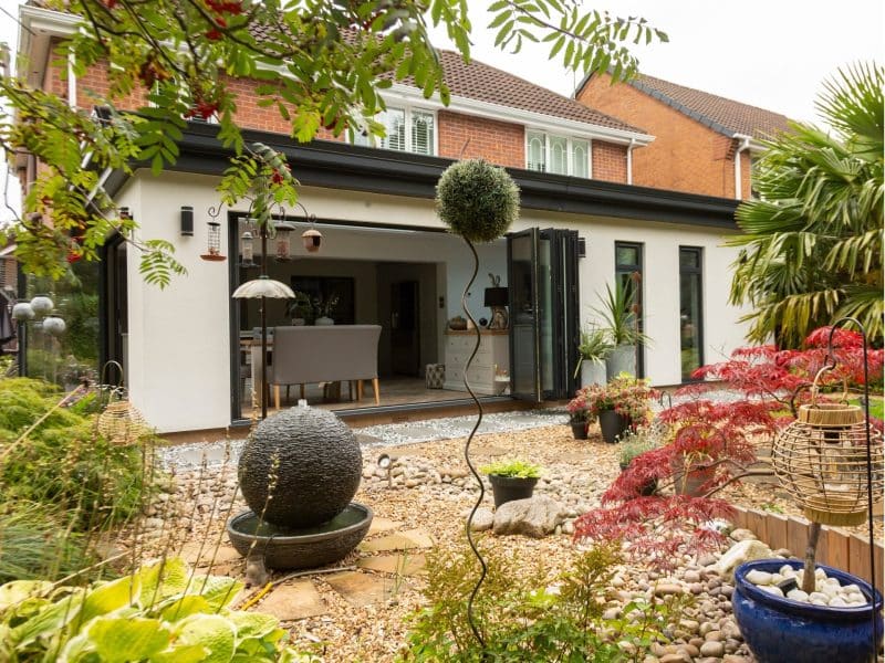 an outside image of a new modular glass extension with cream rendering and grey framed patio doors pulled back showing he new dining area. The patio it leads onto is gravelled with stepping stones and potted plants.